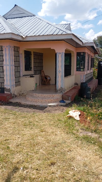 This image depicts a single-story residential building located in Mosop ward, Kenya, under a partly cloudy blue sky. The building features a multi-gabled roof constructed from grey corrugated metal sheets, with white fascia boards and a white PVC gutter system visible on the right side. The exterior walls are painted a light peach-pink, with interspersed sections of exposed grey cinder blocks and decorative vertical bands in alternating light blue and peach-pink on the structural columns and wall surfaces.

A central recessed porch area is accessed by three curved steps, which are tiled with a brown and beige patterned design. The porch floor is similarly tiled. Inside the porch, a wooden rocking chair is positioned, along with a dark-colored door and a small green plastic container. A pair of blue flip-flops rest on the tiled steps. Dark-framed windows are present on the front and right facades of the building. The right side of the structure sits on a raised, dark red-painted concrete foundation, next to which two dark plastic water storage tanks are located.

The surrounding ground consists of sparse, dry yellowish-brown grass interspersed with patches of exposed soil. In the lower right foreground, a light-colored sheet-like object lies on a small mound of reddish-brown earth. No individuals are present in the scene.