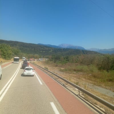 The image shows a highway scene in Champagnier, France. The road is asphalt with white lane markings and a red-colored shoulder. A silver van is partially visible on the left edge of the frame. In the distance, there is a traffic jam with several cars and a white bus. The road is lined with a metal guardrail. Beyond the road, the landscape consists of dry grass and sparse vegetation, with a forested hillside in the background. Mountains, some with snow-capped peaks, are visible on the horizon under a clear blue sky. A power line stretches across the upper right portion of the image.