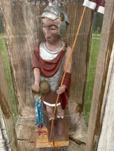 A carved wooden statue of a figure resembling a Roman soldier stands in a display niche. The figure is painted with a brown face, silver armor, a red draped garment, and a brown skirt. It holds a wooden staff with a white and red flag at the top in its right hand and a jug from which blue liquid is being poured in its left hand. At the base of the statue is a small, colorful depiction of a building with red and blue details. The statue is positioned against a weathered wooden background. The display niche is framed by rough-hewn wooden posts, and a reflective surface, possibly glass, is visible in front of the statue, showing the surrounding environment as a blurred green landscape with white lines, suggesting a field. The overall impression is that of an outdoor display, likely in a public space within Sielachowskie, Poland.