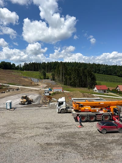 The image depicts a construction site in Jodłownik, Poland, under a partly cloudy blue sky. In the foreground, a large orange crane truck with the "olstál" logo is parked on a gravel surface. Adjacent to the crane truck is a red car with a black roof. To the left of the crane truck, a white truck is partially visible. Further left, a yellow bulldozer is positioned near a portable toilet. In the mid-ground, there are stacks of construction materials, including rebar. Behind these, a small blue portable toilet stands. A temporary structure, possibly an office or storage unit, is also present. The background shows a sloping, cleared area of land, with a dense forest of evergreen trees bordering the site. In the distance, a few residential buildings with red roofs are visible nestled among the trees and vineyards. The overall scene suggests ongoing construction or land development activities.