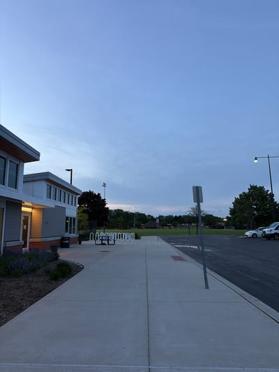 The image depicts a late-day or early-evening scene in what appears to be a park or recreational area in Morton Grove, Illinois, United States.
The foreground shows a modern building with gray siding and multiple windows. Adjacent to the building is a landscaped area with lavender plants and mulch. A sidewalk runs along the building, and a bike rack is positioned nearby. A garbage can labeled "Parks" is also visible.
Beyond the building and sidewalk is a grassy field with trees in the background. Utility poles and possible sports field lights are discernible in the distance.
To the right of the frame, a paved road runs parallel to the sidewalk. Vehicles are parked along the edge of the road. The sky is blue with some clouds, suggesting either early morning or late evening lighting conditions.
