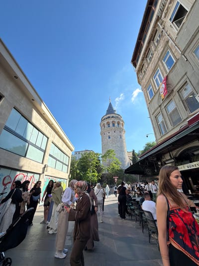 In the city of Beyoğlu, Turkey, a tall, conical-roofed stone tower, likely the Galata Tower, dominates the background under a clear blue sky. To its right, a weathered stone building features arched windows and a red and yellow flag. In the foreground, a bustling street scene unfolds. On the left, a modern building with large glass windows and graffiti lines the street. A diverse group of people, some wearing headscarves, are walking and conversing. A baby stroller is visible on the far left. On the right, a cafe or restaurant with outdoor seating is set up under a dark awning, where patrons are seated at tables. A woman with long brown hair, wearing a dark top with a red floral pattern, is positioned in the immediate foreground, facing away from the camera and looking to the right. The street is paved with stone and illuminated by bright sunlight, casting shadows.