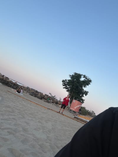 The image depicts a beach scene in Sveti Vlas, Bulgaria, likely during late afternoon or early evening, as indicated by the soft, gradient lighting of the sky. In the foreground, the sandy beach is visible, with a dark, out-of-focus object, possibly clothing or a blanket, obscuring the bottom right portion of the frame.
Further back, on the sandy expanse, two individuals are seated on a blanket near a rocky embankment. One person appears to have dark hair, and the other is wearing a light-colored garment. Beyond them, a young person wearing a red jacket and dark shorts is standing on the sand. To their right, a large tree with dense foliage stands prominently. In front of the tree, a red and white striped banner with text in Cyrillic script, including "РЕЗЕРВИРАЙ МЕ," is displayed, accompanied by a QR code. The rocky embankment runs along the left side of the frame, separating the beach from the sea, where a faint cityscape can be observed in the distance. Wooden planks are laid out on the sand, potentially forming a pathway or part of a seating area.