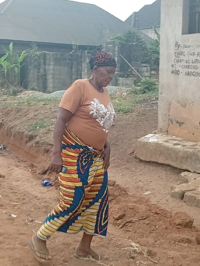 An elderly woman wearing a patterned headscarf, an orange t-shirt with a white floral design, and a long, colorful wrapped skirt walks outdoors. She wears simple flip-flops and is on a dirt path. Behind her are buildings and greenery. To the right is a wall with handwritten words, possibly advertising charging services with prices like "#30 CHARGING." The scene is outdoors, possibly in a rural setting.