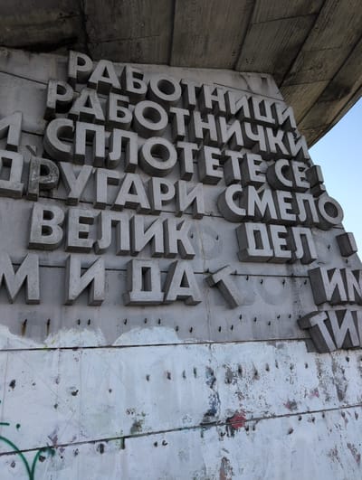 This media file shows a close-up, low-angle view of a large concrete monument with raised, three-dimensional Cyrillic letters. The monument appears to be part of an outdoor structure, possibly a wall or building, made of rough concrete. The letters are arranged vertically and horizontally, forming words and phrases. The sun is high in the sky, casting shadows on the letters and highlighting their texture. The background shows a clear, light blue sky and a section of a rough, textured concrete wall to the left of the monument. The right side of the monument is attached to a white concrete surface that shows signs of weathering, peeling paint, and graffiti, including splotches of color and green spray-painted lines. Small metal fasteners are visible embedded in the white concrete surface. The overall impression is of a monumental, possibly historical, structure in an outdoor setting.
The visible text on the monument, in Cyrillic, reads: "РАБОТНИЧКИ", "РАБОТНИЧКЕ", "РАБОТНИЦИ", "РАБОТИ", "ПРОЛЕ", "ТЕТЕ", "ОТЕ", "СМЕЛО", "ДЕЛ", "ИМ", "ВЕЛИК", "МЕДИ", "МИ". Some letters are partially obscured or difficult to decipher. Based on the location context provided, this is likely the monument to the Bulgarian Communist Party, located on Buzludzha Peak in Bulgaria. The monument is designed to be a powerful symbol of the communist era. The bright, clear sky suggests it is daytime, likely late morning or early afternoon. There are no people visible in the frame.