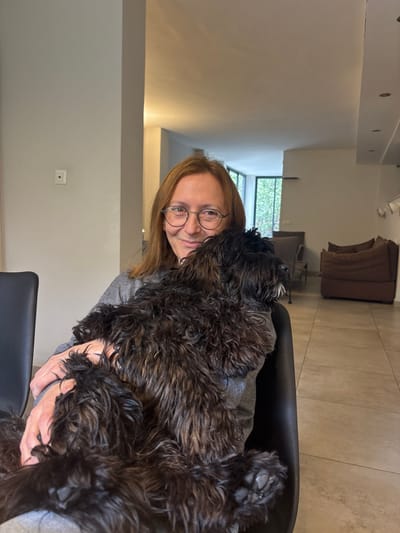 An adult female is seated indoors in what appears to be a residential setting within the city of Ramat HaSharon, Israel. She has medium-length reddish-brown hair, wears round eyeglasses, and a grey long-sleeved top. She is smiling directly at the camera while holding a small to medium-sized dog with shaggy, dark brown fur, which is resting in her arms or lap, facing right and away from the camera. The background reveals a modern interior with light-colored tiled flooring and off-white walls. To the left, a small red square object is mounted on a wall. Towards the right, a large window or glass door displays green foliage outside, and a dark brown sofa or sectional is visible against a wall further back. Recessed lighting is present on the ceiling. The overall scene depicts a relaxed interaction between a person and a pet within a home environment.