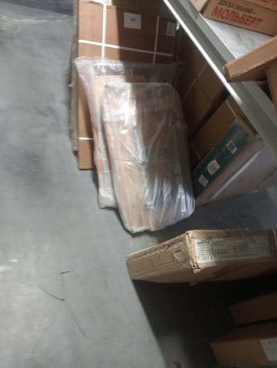 The image depicts a warehouse setting in Aktobe, Kazakhstan, with stacks of cardboard boxes. One prominent box, on the upper right, displays Cyrillic lettering, possibly indicating a brand or product name. The boxes are mostly brown cardboard, some wrapped in clear plastic. The floor appears to be concrete, and there are some metal shelving units visible. The lighting is subdued, suggesting an indoor environment. No people are present in the image.