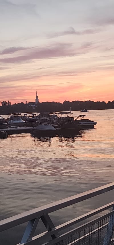 This is a photograph of a marina at sunset, taken from a pier. The sky is filled with streaks of pink, orange, and purple clouds, indicating the sun is low on the horizon. Several boats, including speedboats and pontoons, are docked in the foreground. The water reflects the colors of the sky and the boats. In the background, a treeline separates the water from a building with a tall steeple, possibly a church. The overall scene is tranquil and picturesque. The location is the Chambly marina, in Chambly, Canada.