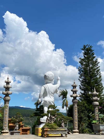 This media file depicts an outdoor scene in Thôn 5, Vietnam, featuring a large white statue of a figure, possibly a child or young person, in mid-stride. The statue is made of smooth, white material and appears to be a prominent element in a garden or temple setting.
In the background, lush green trees and foliage suggest a mountainous or hilly landscape. Several ornate stone structures, possibly pagodas or decorative pillars, are visible, some adorned with intricate carvings and text in a golden hue. The sky is a vibrant blue with scattered white, fluffy clouds, indicating fair weather.
The composition is vertical, with the statue taking up a significant portion of the frame. The statue's pose, with one arm extended, suggests movement or an action being performed. The overall ambiance is serene and spiritual, characteristic of a temple or garden designed for contemplation. The time of day appears to be daytime, likely late morning or afternoon, given the brightness of the sky and the sunlight. The location cues, such as the style of architecture and the lush vegetation, are consistent with a tropical or subtropical setting in Southeast Asia.
The image captures a blend of sculpted art, natural beauty, and architectural elements, creating a visually engaging and peaceful scene.