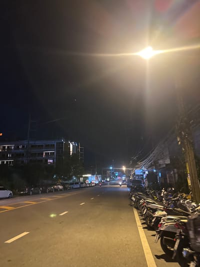 The image depicts a street scene at night in Wichit, Thailand. A bright street lamp illuminates the left side of the frame, causing a lens flare. The street is lined with numerous motorcycles parked along the side. In the background, buildings are visible, some appearing to be under construction. The road has clear lane markings, indicating a well-maintained street. The overall impression is a quiet, ordinary night in a busy urban area. There is no discernible activity or event taking place, other than the general ambiance of a street at night. The lighting suggests it is after dark, and there are no obvious weather indicators. The presence of many motorcycles implies a location where they are a common mode of transportation.
