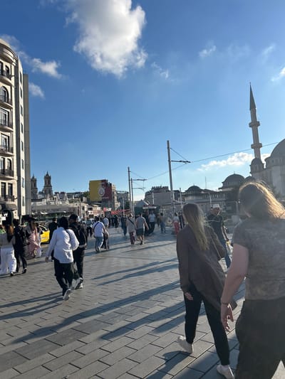 The image depicts a street scene in Beyoğlu, Turkey, under a bright, partly cloudy sky. A diverse group of people are walking on a wide, paved pedestrian walkway. Many are walking towards the viewer, while others are moving away. Some individuals are in the foreground, with their backs or sides to the camera.

Notable elements in the background include modern buildings on the left, with balconies and multiple windows. Further down the street, a large yellow billboard displays the Galatasaray S.K. logo and the text "WE ARE THE CHAMPIONS." A minaret of a mosque is visible to the right, along with dome structures of a mosque complex. A yellow taxi is parked on the street to the left. Overhead wires and street lights are present, suggesting an urban environment with public transportation or street infrastructure. The ground is paved with large, rectangular grey tiles laid in a herringbone pattern, casting long shadows from the people and structures. A person in a wheelchair is also visible among the pedestrians. The overall atmosphere suggests a busy, public urban space during daylight.