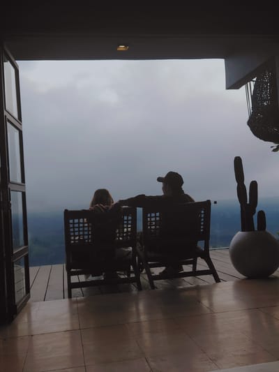 In Kintamani, Indonesia, two individuals are seated in chairs on a wooden deck, facing away from the camera and towards a hazy, expansive view. The person on the left, with long, light-colored hair, is wearing a reddish garment. The person on the right, wearing a baseball cap, has their arm resting on the back of the other person's chair. A large, round planter containing a cactus is positioned to the right of the chairs, and a decorative woven object hangs from the ceiling above. The scene is framed by an open doorway on the left and a tiled floor in the foreground. The sky is overcast, creating a muted, atmospheric backdrop.