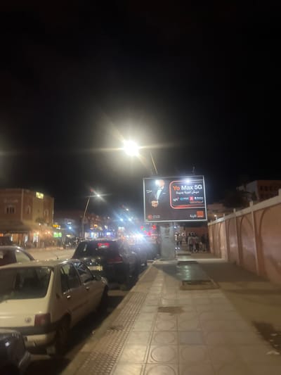 This is a nighttime, low-light wide shot of a busy urban street in Beni Mellal, Morocco. The scene is illuminated by multiple artificial light sources, including bright streetlights, vehicle headlights, and an illuminated billboard.
In the immediate foreground, a light-colored hatchback car (possibly a Peugeot 205) is parked on the left side, facing away from the viewer. Behind it, a row of darker cars is parked along the curb. The asphalt road surface, reflecting some of the ambient light, separates the parked cars from the main flow of traffic, which is visible in the middle distance with numerous vehicle lights.
On the right side of the image, a paved sidewalk extends from the foreground into the background. It is constructed from square tiles, some featuring circular patterns, and includes a tactile paving strip near the curb. A prominent, vertically oriented, illuminated billboard stands on the sidewalk, advertising "Yo Max 5G" with an image of a man in a tuxedo and Arabic text that translates to "Live a new experience," along with "Orange" branding. Beyond the billboard, a group of approximately five to seven pedestrians can be seen walking along the sidewalk.
Further right, a reddish-orange wall with arched recesses runs parallel to the sidewalk, indicating a boundary to a property or structure. In the background on the left, multi-story buildings with reddish-brown facades and visible windows line the street. The sky is dark, indicating a clear night. The overall atmosphere suggests an active commercial or residential thoroughfare.