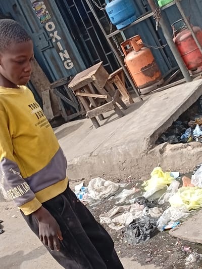 An outdoor urban scene in Sabon Gari West, Nigeria, captures a young male in the left foreground looking downwards. He wears a yellow and grey long-sleeved shirt and black track pants. In the background, a blue corrugated metal structure, possibly a storefront, displays vertical white lettering partially visible as "OKING." Adjacent to this structure, a metal rack holds three distinct propane gas cylinders: a blue one on an upper shelf, an orange, rusted cylinder secured by a chain below it, and a red cylinder partially visible on the far right, also chained. Several crude wooden stools or low tables are discarded on the ground near the blue structure. A raised concrete slab or curb defines the right side of the mid-ground, bordering a ditch overflowing with assorted refuse, predominantly white, yellow, and black plastic bags, indicating an accumulation of waste. The ground comprises dirt and worn concrete. No specific actions or interactions are observed.