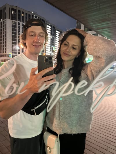 A photograph captures a reflection of a man and a woman in a glass surface at night in Murino, Russia. The man, positioned on the left, appears to be in his late 20s to early 30s, with light brown hair and a dark baseball cap worn backward. He wears a plain white short-sleeved t-shirt and dark pants, with a black fanny pack strapped across his chest. He holds a dark-colored smartphone with a multi-lens camera in his right hand, pointed towards the viewer, suggesting he is taking the picture. He has a slight smile and looks directly at the camera.

The woman, positioned on the right, appears to be in her late 20s to early 30s, with dark, long hair. She wears a light grey, fuzzy knit cardigan with three visible light-colored buttons and dark pants. A small, light beige or cream-colored shoulder bag with a buckle closure hangs at her side. Her left hand is raised, touching the side of her head near her hair. She also has a slight smile and looks slightly to her right (viewer's left).

The background, visible through the reflection, depicts modern multi-story residential or commercial buildings with numerous illuminated windows, indicative of an urban environment. Pedestrian crossing signs are visible in the distance. The ground below appears to be paved with rectangular bricks or tiles. The scene is illuminated by artificial streetlights and building lights against a dark night sky. White script, spelling "Супер" (Super) in Cyrillic, is visibly reflected on the glass surface, partially overlapping the figures. An overhead wooden or textured structure is reflected at the top of the image.