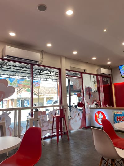 The image shows the interior of a restaurant or cafe in Kecamatan Piyungan, Indonesia (latitude: -7.837, longitude: 110.474). The cafe has a modern design with white walls, a tiled floor, and large windows that offer a view of the street outside. The ceiling has several recessed light fixtures, and a single air conditioner unit is mounted on the wall.
There are several red metal chairs and white tables placed throughout the cafe. The walls are adorned with colorful cartoon-like characters, some of which are holding ice cream cones. Behind the counter, a large sign reads "Mixue HQ," indicating the cafe's brand affiliation. The scene appears to be empty of patrons or staff, suggesting it may be closed or captured during a non-peak period.