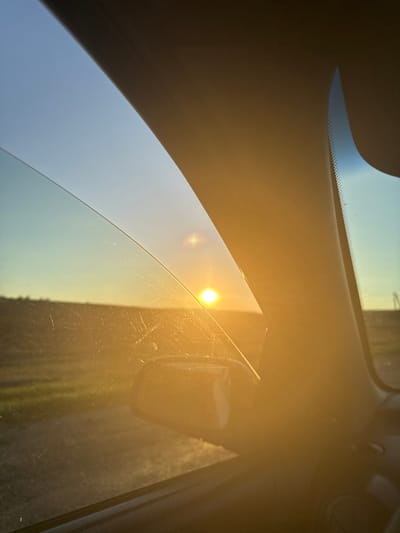 The image is a photograph taken from inside a vehicle, looking out the side window. The sun is low in the sky, casting a warm, golden glow across the scene. It appears to be either sunrise or sunset. The window glass is slightly smudged and scratched.
Through the window, a rural landscape is visible. A field stretches out, with some sparse vegetation and what looks like a dirt road or path. In the distance, a faint line of trees or structures can be discerned. The sky is a clear gradient of blue and yellow, transitioning from a pale blue at the top to a bright yellow where it meets the horizon.
The interior of the car is partially visible, including a portion of the dashboard and a side mirror. The prominent pillar of the car's interior frames the view, obscuring a portion of the landscape and sky.
The specific location in Iordanovca, Moldova, cannot be determined from the visual information presented in the image alone. The scene depicts a generic rural setting consistent with many such locations. There are no discernible landmarks or identifying features that would pinpoint the exact location within Iordanovca.