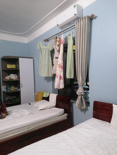 The image captures a brightly lit bedroom or guest room, likely in Bai Chay Ward, Vietnam. The walls are painted a light blue, contrasting with a white ceiling featuring decorative molding.
In the foreground, two beds with wooden headboards and white quilted bedding are visible. On the bed to the left, a person is partially visible, wearing a red shirt and dark cap, looking down at a phone or tablet. A remote control rests on the white comforter. The other bed on the right is empty except for a white pillow and some folded clothes (a yellow t-shirt, a green cloth, and a white towel) placed near its headboard.
Against the back wall, a tall, narrow cupboard with a dark wooden frame and a white sliding door stands to the left. Its open shelves are filled with various items, including folded clothes, plastic bottles, and bags. Above the window, an air conditioner is mounted, displaying the "Gree" logo, "R410A," and a green energy efficiency sticker.
A metal curtain rod extends across the window, holding several items of clothing on hangers, including a light green gingham dress, a polka-dot dress, a floral dress, and a solid light green dress. A silvery-beige curtain, tied in the middle, covers part of the window. Through the gaps in the curtain and above the clothes, decorative window elements with blue backgrounds and yellow floral patterns are visible, suggesting either a patterned window film or a view of a colorful building detail outside.
The room appears tidy, though lived-in, with items stored on shelves and clothes hanging to dry or be aired out. The lighting suggests it is daytime.