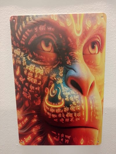 The image is a close-up, head-on view of a metal print artwork featuring the face of Hanuman, a deity from Hindu mythology, depicted in a highly stylized and colorful manner. The artwork is mounted on a textured, light-colored wall, likely concrete or stucco, which is illuminated by a soft, diffused light. The artwork itself is dominated by warm tones of orange, red, and yellow, with accents of blue and green on Hanuman's face. Intricate patterns and what appear to be devotional script in Hindi or Sanskrit are integrated into the artwork, particularly around the eyes and on the lower half of the face. The wall texture is clearly visible around the edges of the print, and the print has two small holes near the top corners, suggesting it is intended to be hung. The print has a slight sheen, indicating a metallic finish. There are no people or visible environmental details of Nuremberg, Germany, present in the image, which focuses solely on the artwork and its immediate background.