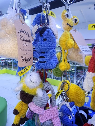 The image shows a display of crocheted keychains hanging from metal hooks. The keychains are of various animals and characters, predominantly in bright colors such as yellow, blue, and white. Some of the keychains are attached to a brown tag that reads "Thank you for celebrating with us." Yellow price tags with barcodes are visible on some items. In the background, shelves with merchandise are visible, suggesting a retail environment. A green object, possibly a stool or seating, is partially visible in the lower left corner. The lighting appears to be artificial, illuminating the display of keychains. There are no people clearly visible in the image, and no specific actions or interactions are depicted. The context implies a store or market in Ushtobe, Kazakhstan, selling these handmade items.