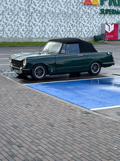 An overhead, eye-level shot captures a dark green Triumph Herald convertible parked in a lot. The car is angled slightly towards the viewer, with its front left wheel closest to the camera. The convertible top is up and appears to be black. The car has chrome trim on the bumpers and along the side, and distinctive wire-spoke wheels. A license plate with the characters "737 AKB" is visible on the front.

The car is parked in a marked parking space, indicated by blue paint and white lines. The ground surrounding the parking space is a grey asphalt, and in the foreground, a cobblestone walkway is visible, with alternating shades of grey and brown bricks laid in a diagonal pattern. The overall scene suggests an outdoor environment, possibly a retail area, with a building facade in the background.

The building facade features a repeating pattern of stylized bird silhouettes in grey against a lighter background. Prominently displayed on the building are the words "SUPERMA" in large green letters, implying a supermarket. To the right of the main building, a red kiosk-like structure with white trim and a red graphic element is visible, possibly related to advertising or information. A shopping cart is partially visible near the building's entrance. The ground appears wet in some areas, suggesting recent rain. The environment suggests a suburban or commercial area.