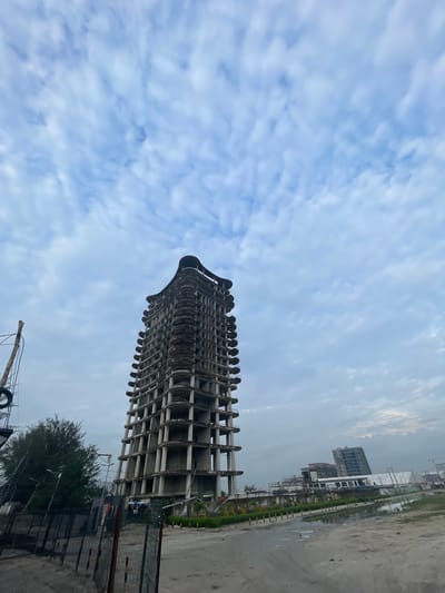 An unfinished, tall, concrete building stands prominently in the foreground of the image. The building has a unique, curved design with multiple balconies or floor extrusions. It appears to be in a state of construction or abandonment, with exposed concrete and a lack of finishing.
To the left of the main building, a bare, weathered wooden pole is visible, possibly a utility pole or part of a structure. A dark fence runs across the bottom left of the frame, with some vegetation behind it.
In the middle ground and background, other buildings are visible, also appearing to be under construction or in a similar unfinished state. These include several multi-story structures and what looks like a long, low-lying building with scaffolding. The environment around the buildings is a mix of sandy ground, some puddles of water, and patchy greenery. The sky above is filled with a pattern of light, wispy clouds against a pale blue background.
The scene is located in the city of Ilado, Nigeria, and captures a developing or stalled construction area within the urban landscape. No people are visible in the image.