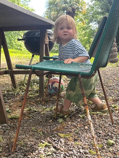 In Erba, Italy, a young child with blond hair, wearing a blue and white striped t-shirt and green camouflage shorts, sits on a weathered green wire chair next to a charcoal grill. The child is looking towards the camera with a neutral expression. A small soccer ball featuring the character Stitch is positioned on the ground beneath the chair. The scene is set outdoors on a gravelly surface scattered with leaves, with a wooden table and lush green foliage in the background, suggesting a garden or park environment. The chair's metal legs are rusty, indicating it has been exposed to the elements.