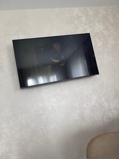 A flat-screen television is mounted on a textured, light-colored wall. The television screen displays a dark image with a person's face visible, appearing to be a still from a movie or show. Reflections of light and possibly curtains are visible on the glossy surface of the screen. The overall scene suggests an indoor setting, likely a room in a residential or hotel property. The location is specified as Nizhnyarkeyevo, Russia.