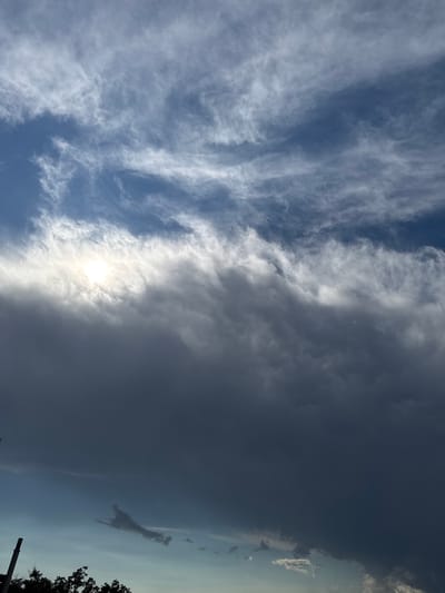 The image captures a dramatic sky over Pelinovo, Montenegro, dominated by a complex interplay of clouds. The upper portion of the frame reveals a bright blue sky streaked with wispy cirrus clouds, illuminated by the sun which is partially obscured by a thick band of cumulonimbus clouds below. These lower clouds are dark and heavy, suggesting an impending storm or the aftermath of one. The transition between the bright, thinner clouds and the dark, dense ones creates a striking contrast. In the lower left of the image, the silhouettes of trees and a pole are visible against the lighter sky, grounding the scene in its geographical context. The overall impression is one of powerful natural forces at play in the sky above the Montenegrin landscape.