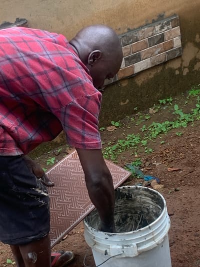 A bald Black man is working outdoors on a muddy ground in Agban, Nigeria. He is wearing a red and black plaid shirt and dark shorts. He is bent over, holding a patterned tile in his left hand and dipping it into a white bucket filled with grey mortar. The background shows a tan wall with a section of decorative tiles affixed to it. There are patches of green vegetation and dirt on the ground.