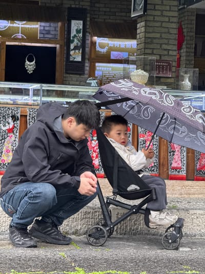 The image captures a man sitting beside a child in a stroller beneath an umbrella. The man is wearing a dark jacket over a gray hooded sweatshirt and blue jeans, with black sneakers on his feet. The child is in the stroller, covered by a gray umbrella patterned with large paisley shapes. The background includes a brick wall and glass display cases with various items. The image was taken in the city of Zhuoshui, China.