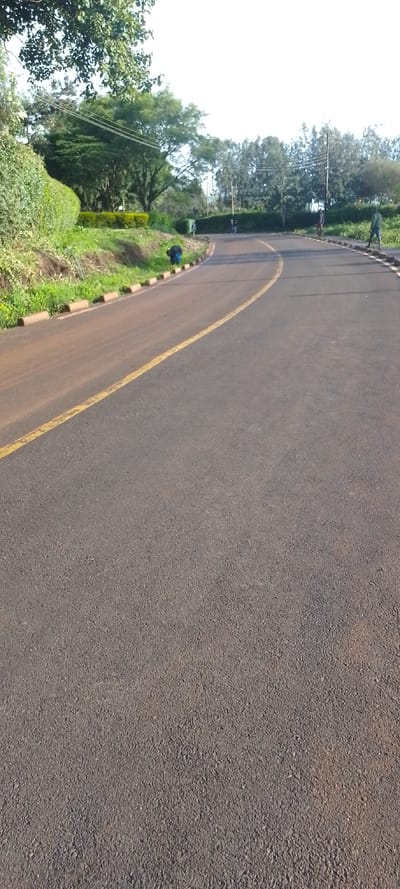 An asphalt road with a solid yellow dividing line extends into the distance, curving right. The road surface is dark grey, with areas of reddish-brown dirt adhering to it. On the left, a raised dirt embankment with green vegetation and small, light-brown concrete curb segments runs alongside the road. A person wearing a blue garment is bent over at the base of this embankment. Further along the left side, a dark green utility box is visible near the road edge. On the right, similar curb segments border a dirt and grass verge, beyond which stands a line of trees. Several individuals are present in the distance on the right side of the road; two are standing or walking on the verge, and one is further back, possibly walking or cycling on the road. Overhead utility lines span across the scene, supported by poles on the right. Lush green trees and bushes dominate the background, particularly on the left, under a bright, clear sky. The scene is located in Ngong ward, Kenya.