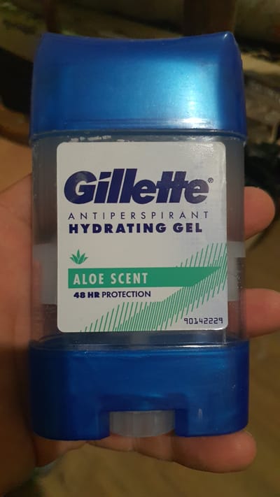 The image shows a close-up of a Gillette antiperspirant hydrating gel with aloe scent, held by a person's hand. The product is in a clear plastic container with a blue cap. The label on the container displays the Gillette logo, "ANTIPERSPIRANT HYDRATING GEL," "ALOE SCENT," and "48 HR PROTECTION." A product code, "90142229," is also visible on the label. The background is blurred and appears to be indoors, with a wooden surface visible. There is no indication of the city of Karankul, Uzbekistan, in the image or its context.