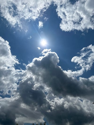 The image displays an expansive daytime sky in Wisła, Poland, featuring a striking contrast between sunlight and varied cloud formations. The bright sun is positioned in the upper left portion of the frame, emitting strong rays and a visible lens flare against a clear, deep blue sky.
Surrounding the sun are clusters of luminous white clouds, appearing soft and fluffy, likely illuminated by the direct sunlight. Moving across the sky towards the right, the clouds transition into much darker, heavier grey formations, with significant shadowing that indicates their depth and density. These darker clouds dominate the right side and upper central areas of the image, creating a dramatic visual division from the brighter, sunlit parts of the sky.
Along the very top right edge of the frame, a few power lines are faintly visible, stretching diagonally across the corner. Additionally, some dark green tree leaves can be discerned on the far right edge, suggesting the photograph was taken looking upwards from ground level, near some natural and man-made elements. There are no people, objects, or text prominently featured, with the primary focus being the dynamic sky and its atmospheric conditions. The scene captures a moment of bright, partially cloudy weather.