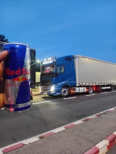 In Sekule, Slovakia, a person's hand is holding a can of Red Bull energy drink in the foreground. The can is blue and silver with red lettering. Behind the can, a blue semi-trailer truck is parked on the side of a road. The truck has a white trailer and is lit up. The sky is a deep blue, indicating it is either early morning or late evening. The road has white lines painted on it, and there is a curb with red and white markings along the side. The overall scene appears to be a roadside stop, possibly at a truck stop or rest area.