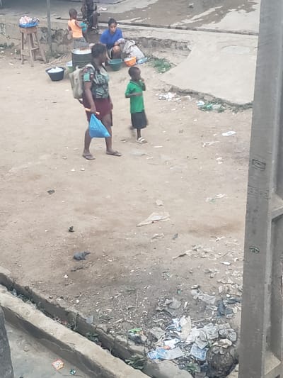 The image depicts an outdoor scene in Alalubosa, Nigeria. Several people are present, including children and adults. Some individuals are lying on the ground, while others are seated around what appears to be a cooking area with pots and containers. The surrounding environment is characterized by dirt and debris. There are some concrete structures visible in the background and along the edges of the scene. The location seems to be an open, unpaved area, potentially near a residential zone.