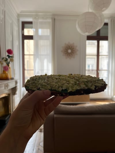 The image depicts a person holding a long, filled pastry, likely an éclair, in front of a window in what appears to be a residential or commercial space. The éclair has a dark pastry base, a green filling, and a generous topping of small, light-colored, irregular pieces. The person's hand and lower arm are visible in the foreground, holding the pastry.
The background reveals details of the interior and exterior of the location. A set of windows with multiple panes is visible behind the éclair. A decorative light fixture comprising several round paper lanterns hangs near the ceiling. The walls are white, and there is a decorative floral design on the wall. In the lower part of the frame, there's a glimpse of a mantelpiece with pink flowers in a vase. Outside the window, details of the urban landscape are visible.
Based on the architectural style and the style of the interior, the image was likely taken in Lyon, France. Lyon is characterized by buildings with tall windows, multiple panes, and ornate interiors.