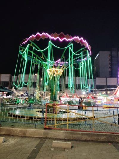 The image shows a green and purple illuminated amusement ride in an outdoor space in the city of Telukjambe, Indonesia, at coordinates -6.306, 107.278. The ride is a circular platform with multiple chains hanging from it. The chains are illuminated with green lights and the platform has purple lights around the edge. The ride is surrounded by a metal fence. Other amusement rides are visible in the background, as well as buildings and a paved sidewalk. Two people are visible in the background standing next to the fence. There is a brick sidewalk in the foreground. The sky is dark and the scene is lit by artificial light.