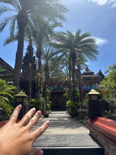 A hand is held up in the foreground, obscuring the view of a pathway leading through a lush courtyard. Tall palm trees flank the path, with their fronds reaching towards a bright blue sky dotted with wispy clouds. In the background, a multi-story building with traditional Thai architecture is visible, featuring dark wood accents and tiered, pointed roofs. The building is surrounded by vibrant green foliage and decorative elements, including golden urns and ornate lanterns. The scene appears to be a serene outdoor area, possibly a temple or a resort in Baan Lamai, Thailand.