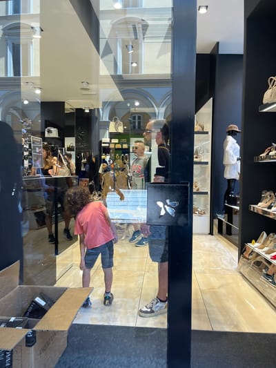 The image shows the storefront of a retail store in Bologna, Italy, reflected in a glass window. Inside, shelves display handbags and shoes. A mannequin stands near the top shelf. The reflections on the glass show two people, likely a child in a pink shirt and an adult, standing outside the store. Several cardboard boxes sit on the ground near the bottom right of the window. The reflection shows architectural details, including arched windows, of buildings across the street.