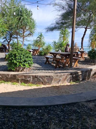 The image depicts an outdoor seating area at what appears to be a beachside cafe or restaurant in Monikeun, Indonesia. The scene is characterized by wooden picnic tables and benches set on a gravel ground, shaded by palm trees and other lush greenery. In the background, the calm blue sea stretches to the horizon under a clear sky, suggesting a pleasant, possibly sunny day.

Several individuals are visible seated at the tables, seemingly enjoying a meal or conversation. One person is wearing a hijab, as is another person further down the seating area, indicating a likely cultural context. The presence of outdoor lighting suggests the area might be used in the evening as well, though the bright sky points to daylight hours. The foreground features a pathway made of stone and sand, bordering a section covered with dark pebbles. Some small plants are growing along the stone wall. The overall atmosphere seems relaxed and serene, a typical beachside dining experience. No visible text is present in the image.