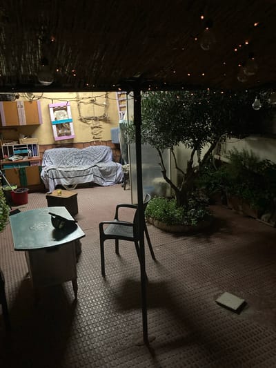 The image depicts a dimly lit outdoor patio area, likely in Sabadell, Spain, at night. A thatched roof covers part of the space, adorned with string lights and a few exposed bulbs. Beneath the roof, a couch draped with a patterned cover faces a wall with shelving and decorative items. To the left, a green metal table with a dark object on it, possibly a bag, stands in the foreground. A black plastic chair is positioned near the table. To the right, a small olive tree grows within a circular planter surrounded by lush greenery. In the background, more plants in planters are visible. The ground is covered in a red, textured material. A square, light-colored tile or object lies on the ground to the right of the frame.