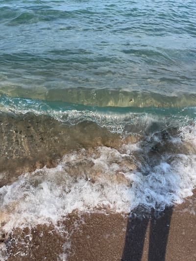 The image shows a close-up view of a wave breaking on a sandy shore. The water is clear and turquoise, with sunlight glinting off the surface. The wave is creating white foam as it rolls onto the beach. The sand is a light brown color and appears wet. In the lower right corner of the image, two dark shadows are cast on the sand, suggesting the presence of people or objects out of frame. The scene is likely a beach in Ahtopol, Bulgaria, given the context.
