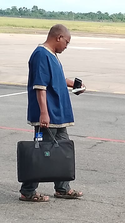 An adult male is standing on an asphalt surface, likely an airport tarmac, in Orerokpe, Nigeria. He is wearing a dark blue tunic with gold trim around the collar and sleeves, dark trousers, and brown sandals. He is bald and wearing glasses. In his right hand, he is holding a smartphone and what appears to be a passport or wallet. His left hand is holding the handle of a large black duffel bag. The bag has a small, light green emblem on its front. In the background, there is a grassy area with trees, and beyond that, a pale sky. The ground is marked with yellow and red lines.