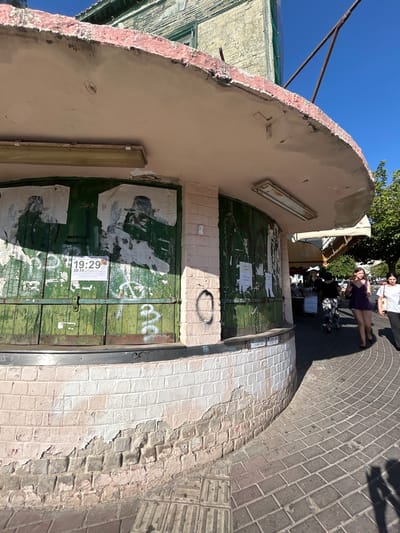 The image depicts an outdoor scene in Rishon LeZion, Israel, under a clear blue sky, suggesting fair weather and likely daytime. The main focus is a weathered, green wooden double door set within an arched entrance. The door is peeling and covered with torn posters, some featuring faint images and graffiti. A flyer with the times "19:29" and "20:34" is prominently attached to the door.
To the right of the entrance, the wall is constructed of bricks, some of which are chipped and have peeling paint, revealing older layers beneath. The pavement is made of cobblestones, curving away from the entrance.
In the background, near the top of the image, several people are visible. Two individuals, one wearing a purple dress and the other a white t-shirt, are standing on the cobblestone path, engaged in conversation. Their body language suggests a relaxed social interaction. Beyond them, a glimpse of market stalls or shops with awnings can be seen, hinting at a commercial or public area.
On the left side of the entrance, part of a building with a textured, light-colored facade is visible, along with some metal structures and netting. The overall impression is of an older, somewhat dilapidated entrance to a building or area, possibly in a historic or developing part of the city. The sunlight casts shadows, indicating it is likely mid-afternoon.