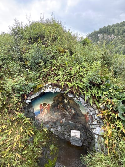 The image depicts a sacred spring located in Chemal, Russia, a small and revered site nestled within a lush, overgrown natural landscape. The spring is housed within a small grotto, its opening framed by rough-hewn stones and abundant green vegetation, including ferns and other leafy plants. Dominating the grotto's interior is a painted mural depicting a religious scene.

The mural features an angel with large, red wings on the left, rendered in a traditional iconographic style with golden halos. To the right of the angel, other figures are depicted, possibly from a Nativity scene, with a star prominent above them. The background of the mural transitions from a starry sky to a rocky landscape.

At the base of the grotto, water pools, appearing dark and slightly murky, reflecting the surrounding greenery and the mural. A small, white sign with red Cyrillic text is affixed to a stone within the grotto, near the water's edge. The text on the sign reads "ДЕНЬГАМИ НЕ СОРИТЬ," which translates to "Do not litter with money," a common directive at religious sites, suggesting that offerings or monetary donations are not to be discarded in the water.

The environment surrounding the grotto is verdant and untamed, with thick foliage climbing up a hillside. In the background, a steep, rocky mountain slope covered in dense forest under a cloudy sky can be seen. The overall impression is one of a tranquil, spiritual place integrated into a wild, natural setting.