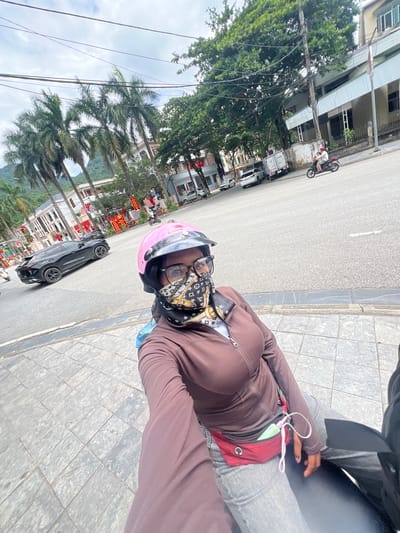 A person wearing a pink helmet, glasses, and a brown jacket is sitting on a scooter, facing the camera. They have a red fanny pack around their waist, with a phone and earbuds visible. The scooter is on a paved sidewalk next to a street. Across the street, there are buildings, palm trees, and a black SUV. In the background, there are more trees, buildings, and a cloudy sky. The image appears to have been taken in the city of Hà Giang 2 Ward, Vietnam.