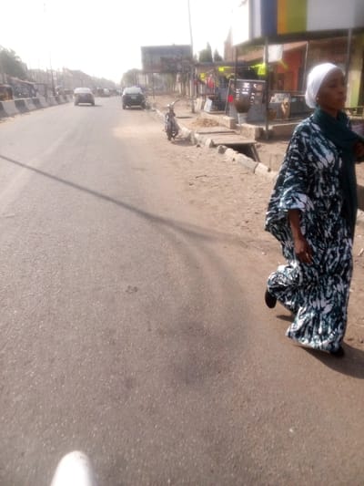 The image captures a street scene in Sabon Gari West, Nigeria, under bright, clear daylight. The main subject is a woman walking on the right side of a paved road, moving away from the viewer. She is dressed in a long, loose-fitting kaftan with a white and dark green/blue pattern, a white head covering, and a dark green scarf. Her dark shoes are visible.

The road is asphalt, appearing well-worn, and has a dirt shoulder on the right. A concrete curb and an open drainage channel separate the road from the unpaved area. Along the dirt shoulder, a motorcycle is parked, and a series of makeshift market stalls or small shops with varied roofing (some corrugated iron) line the street. Further down, more permanent buildings are visible, along with a large billboard that has some illegible text. In the distance on the road, two cars are visible, one red and one dark, suggesting light traffic or parked vehicles.

The bright sunlight casts distinct, moderately long shadows on the road, particularly from a utility pole or similar structure and the walking woman, indicating it is likely late morning or early afternoon. The sky is bright and hazy. No clear emotions are discernible from the woman’s side profile. A white object, possibly the photographer's shoe, is partially visible in the bottom left corner of the frame. No legible text is present on any signs or billboards.