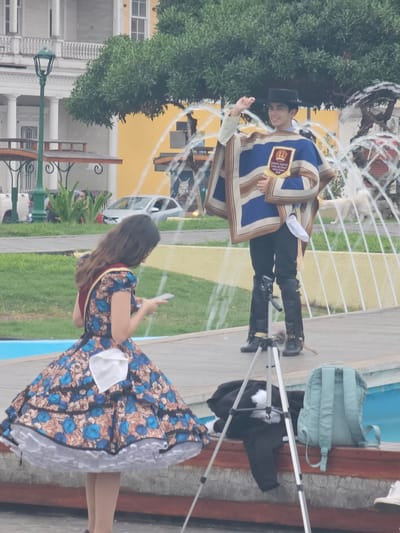 An outdoor scene in Iquique, Chile features two people in traditional attire. A young man, facing the viewer and smiling, wears a black hat, a blue and tan poncho with a crest, and black pants tucked into fringed boots. He is gesturing with his left hand. To the left and in the foreground, a young woman with dark hair is turned away from the camera, looking down at her phone. She wears a full, floral-patterned dress with a red sash and a white kerchief tucked into the waist. Behind them, a fountain sprays water, and lush green trees are visible. A building with classical architectural elements and a bright yellow wall forms the backdrop. In the lower right portion of the frame, a light blue backpack rests on a wooden bench next to a tripod holding a camera.