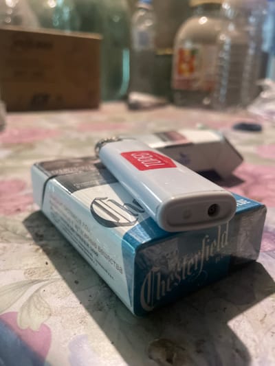 The image depicts a pack of Chesterfield cigarettes with a white lighter on top. The setting appears to be indoors, possibly a residence, in Olginka, Russia.